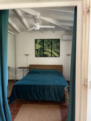 Un dormitorio con una cama, un ventilador de techo, aire acondicionado en la pared, una mesita pequeña y una cesta tejida.