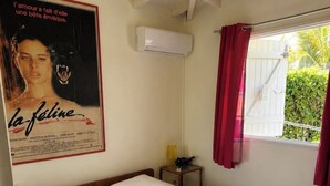 A bedroom with a single bed, a ceiling fan, an air conditioning unit, a window with red curtains, and a poster on the wall. - Villa Sainte-Anne (Sainte-Anne)