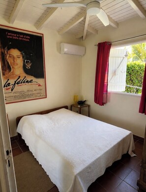 A bedroom with a single bed, a ceiling fan, an air conditioning unit, a window with red curtains, and a poster on the wall. - Villa Sainte-Anne (Sainte-Anne)