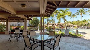 Une terrasse extérieure avec une table et des chaises en verre, entourée de palmiers et d’une piscine.