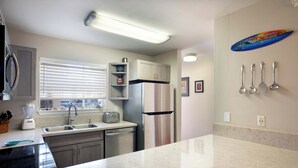A modern kitchen with a stainless steel refrigerator, a built-in oven, a toaster, and a blender. There is a window with blinds, a countertop with a knife block, and a wall-mounted knife rack.