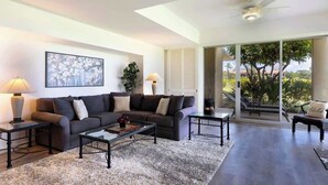 Un salon moderne avec un canapé, une table basse et des lampes. Une grande fenêtre offre une vue sur les arbres, et il y a un balcon.