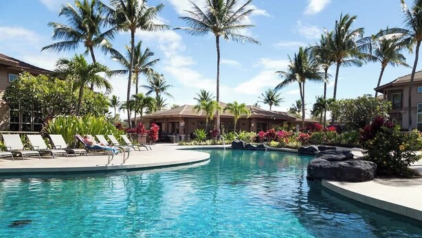 A pool area with lounge chairs, palm trees, and a building in the background. - Pools/Hot tub, BBQ area, Beach gear, Views, Tennis (Waikoloa Village)