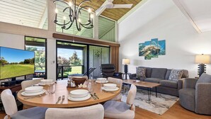 A spacious living room with a large TV, a dining area with a wooden table set for four, and a grey sofa. - Newly Remodeled!! 2BR, Pools & hot tubs, Golf View (Waikoloa Village)