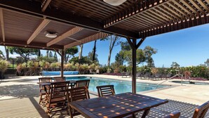 A covered outdoor area with a pool, wooden furniture, and a view of trees and a clear sky. - Newly Remodeled!! 2BR, Pools & hot tubs, Golf View (Waikoloa Village)