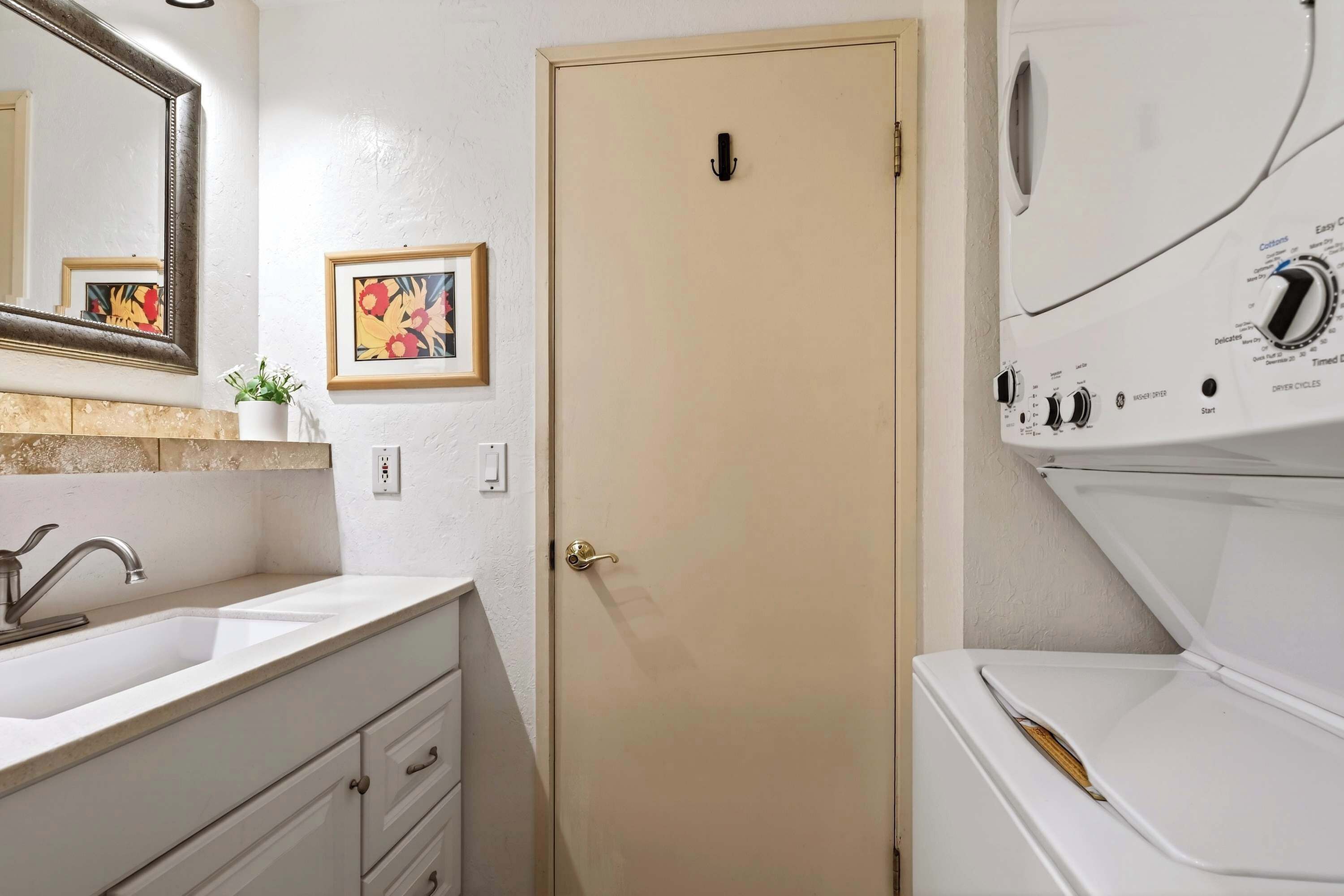 A bathroom with a sink, mirror, framed artwork, and a door leading to a laundry area with a washing machine.