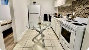 Una cocina compacta con gabinetes blancos, una mesa con cubierta de vidrio, un refrigerador blanco, una estufa y un salpicadero de azulejos.