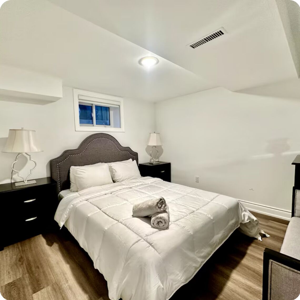 Une chambre à coucher avec un lit, deux tables de chevet, une fenêtre et une bouche d’aération au plafond.