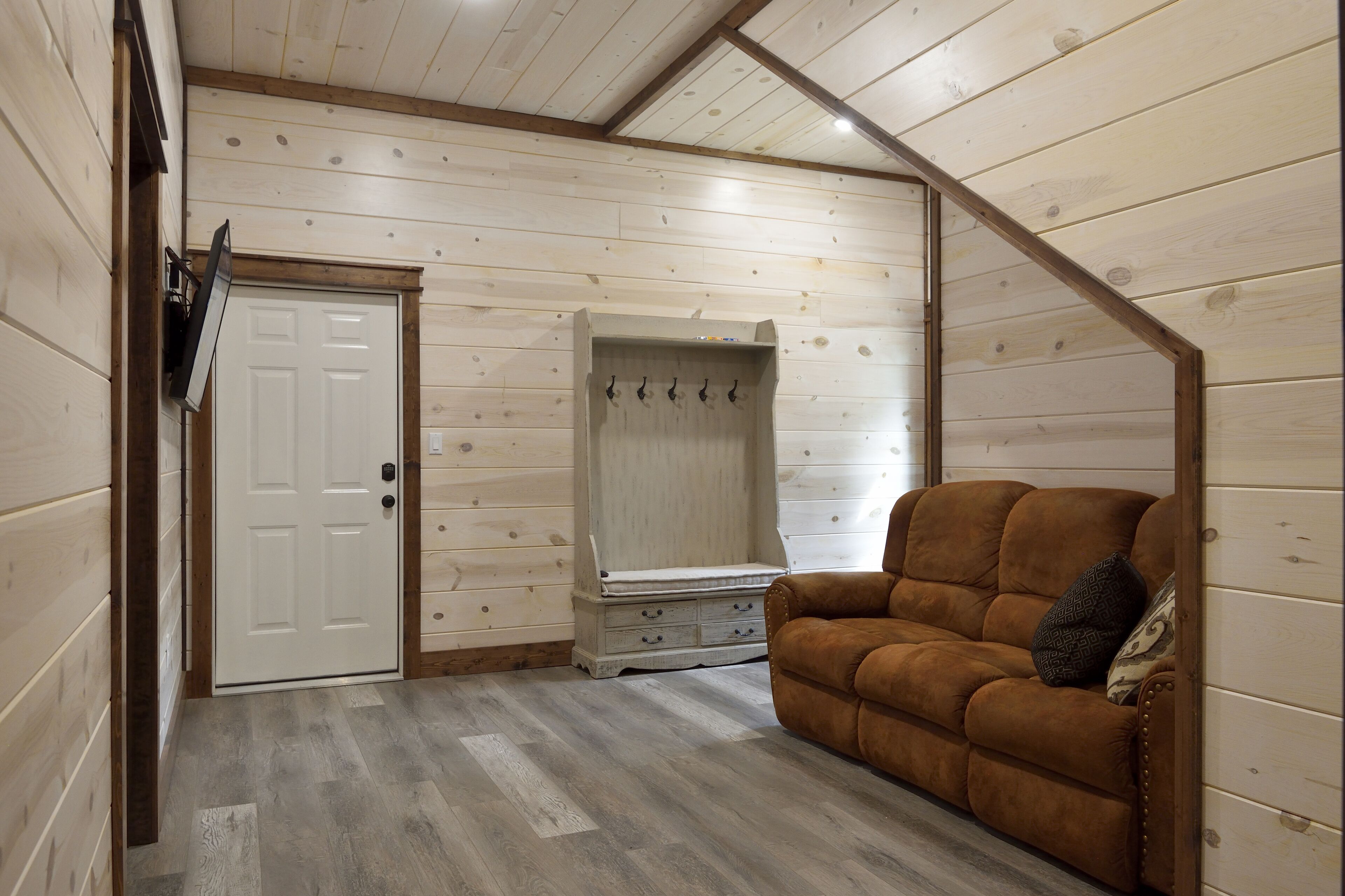Una habitación con paredes de madera, una puerta blanca, un perchero, un sofá marrón y un pequeño aparador.