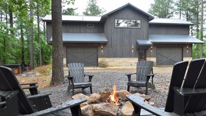 A rustic cabin with a fire pit surrounded by Adirondack chairs, set in a wooded area.