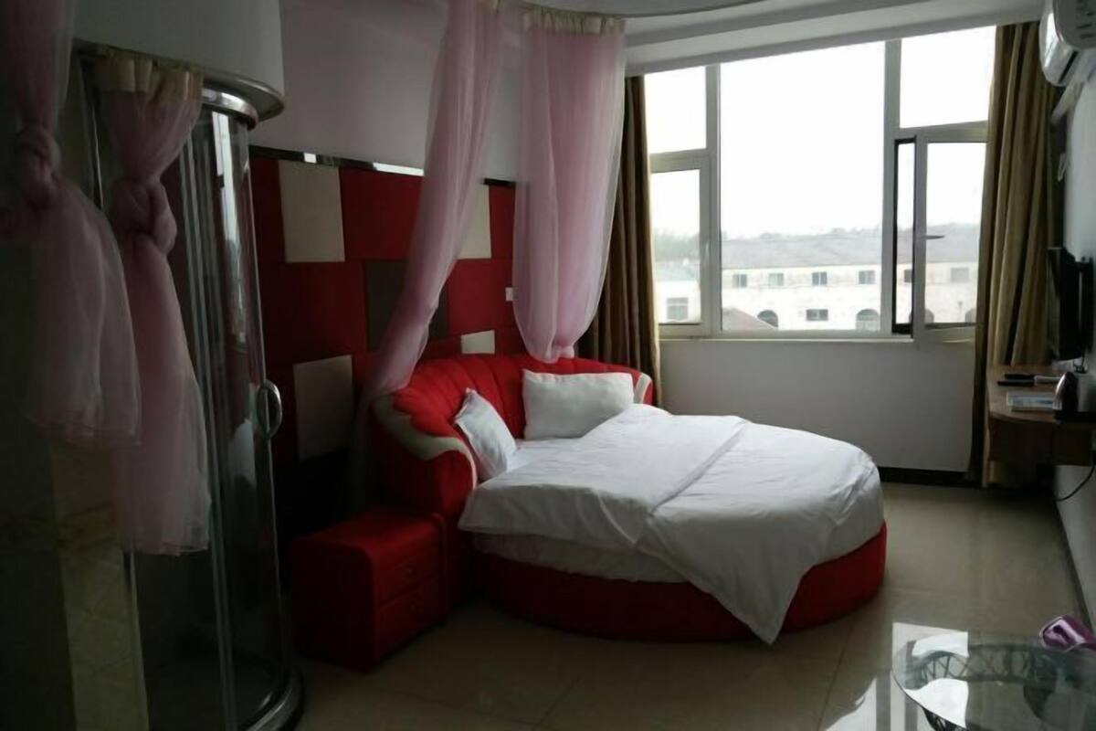 A bedroom with a red bed, white bedding, a glass shower enclosure, and a view of buildings through a large window.