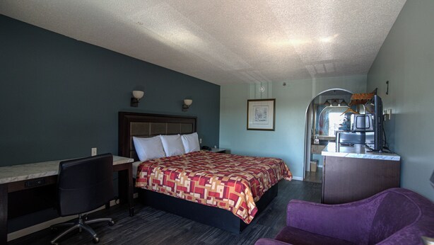 A hotel room with a bed, desk, chair, and a kitchenette area. - Excel Inn & Suites (Fredericksburg)