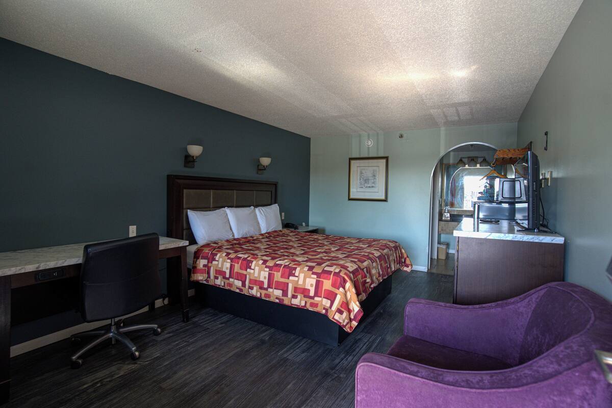 A hotel room with a bed, desk, chair, and a kitchenette area.
