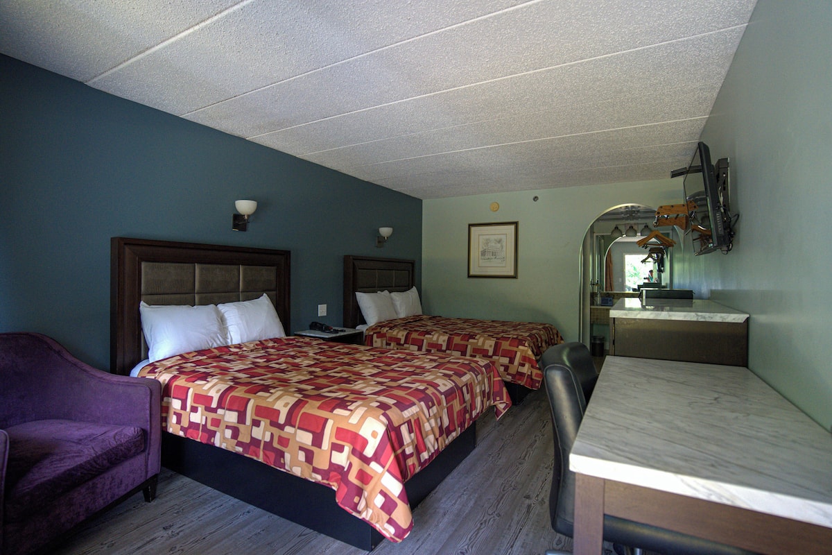 A hotel room with two beds, a desk, a chair, and a bathroom visible through an archway.
