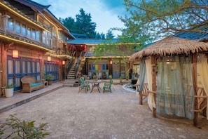 Une cour bordée de bâtiments traditionnels en bois, une structure à toit de chaume et des espaces de repos en plein air.