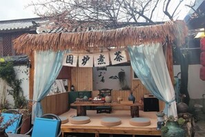 An outdoor seating area with a thatched roof, wooden furniture, and a sign with Chinese characters. - Lijiang Shu He Shangshanruoshui Inn (Lijiang)