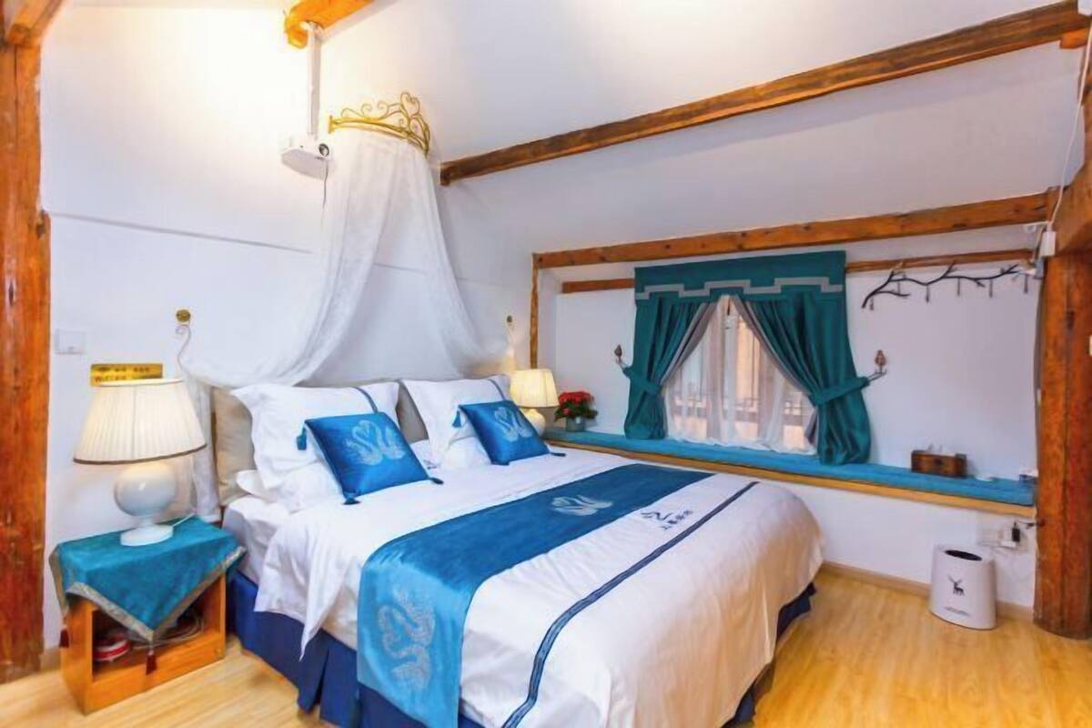 A bedroom with a bed, a bedside table, a lamp, a window with curtains, and a wooden ceiling beam.