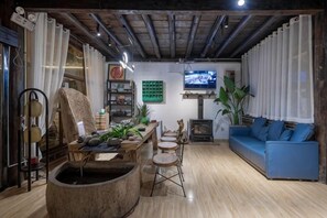 A room with a wooden floor, a blue sofa, a wooden table with plants, a TV mounted on the wall, and a large window with curtains. - Lijiang Shu He Shangshanruoshui Inn (Lijiang)