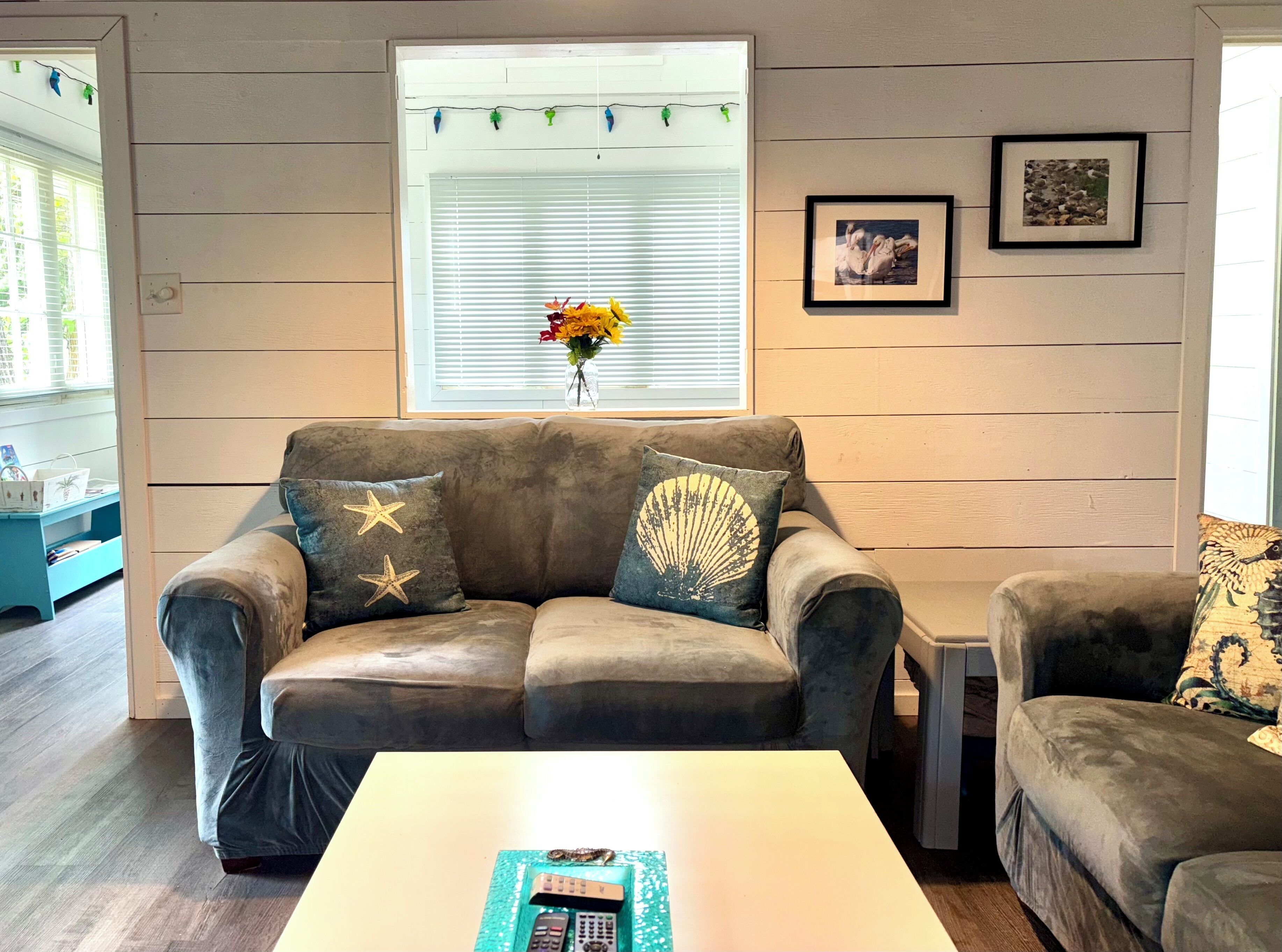 A living room with a brown leather sofa, a white coffee table, and a small side table. There is a window with blinds and a picture frame on the wall.