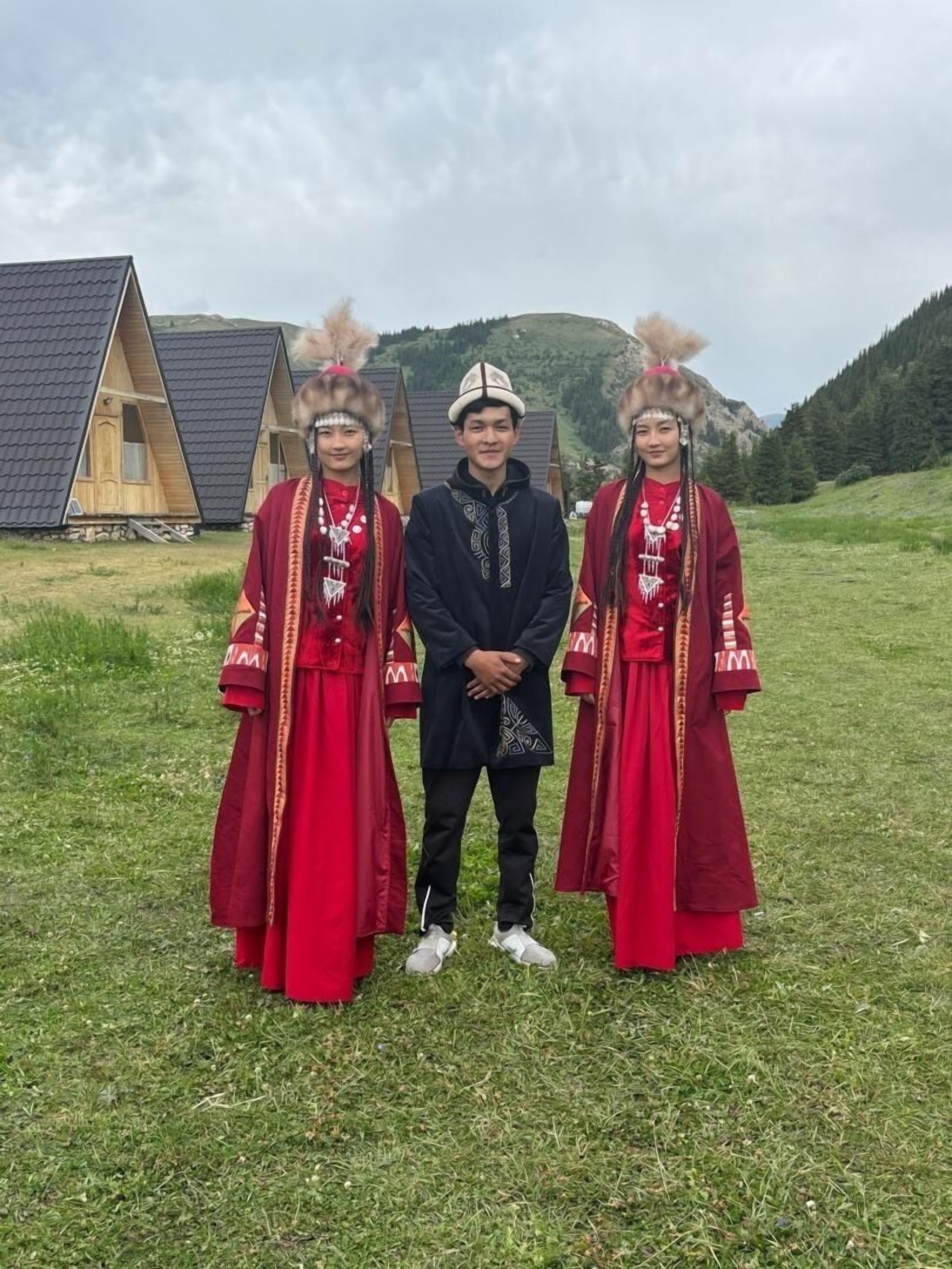 Tres personas vestidas con traje tradicional de pie en un campo con cabañas de madera al fondo.