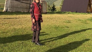 A person standing in front of a traditional yurt with a flag, a rainbow, and a modern building in a grassy field. - Bosogo Resort (At-Bashy)
