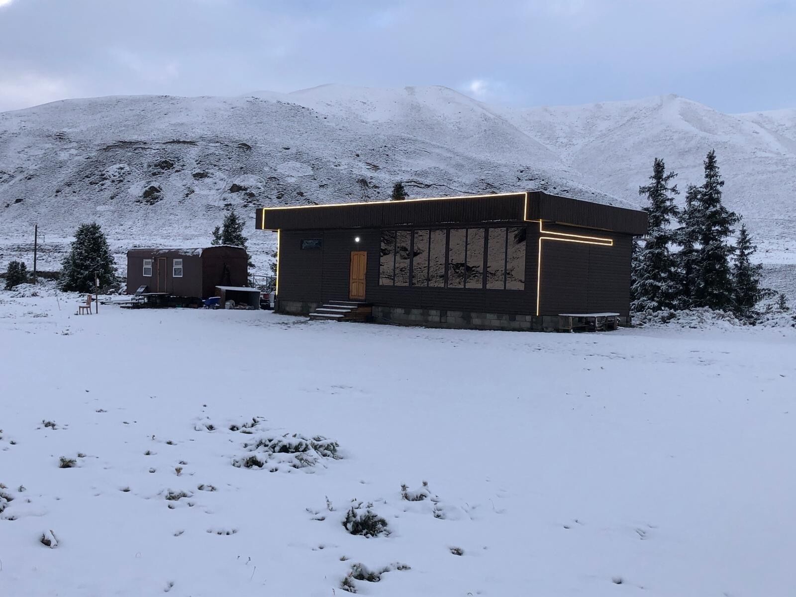 A cabin with a large window and a door, surrounded by snow-covered ground and trees, with mountains in the background.