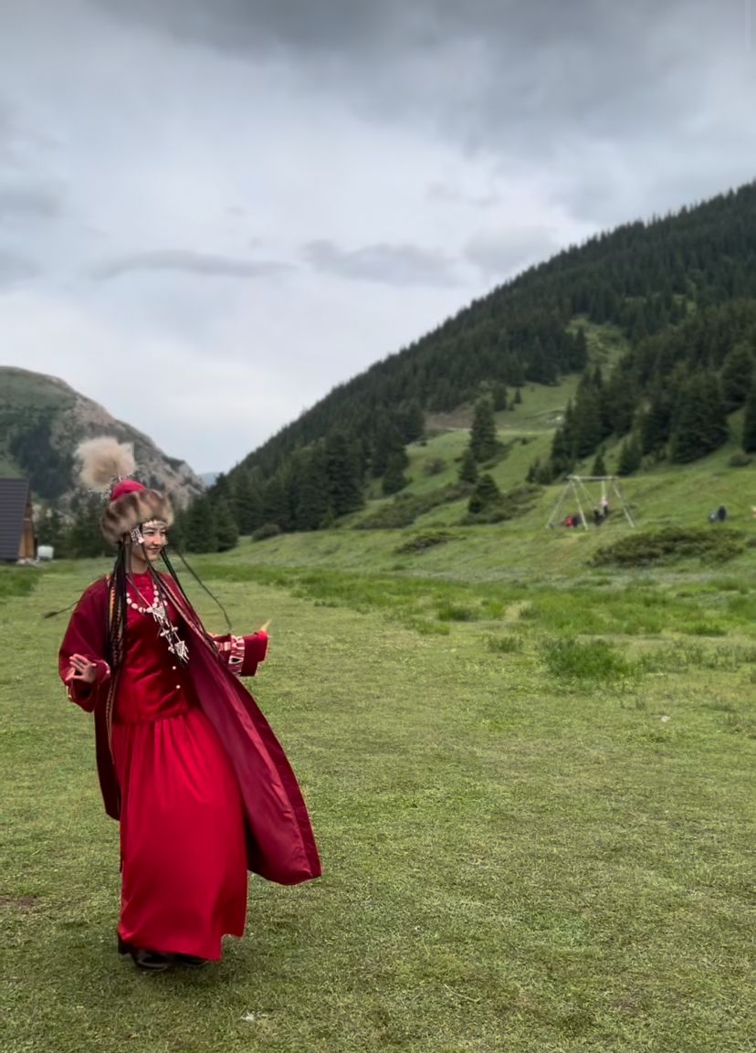 A person in a red dress walking in a grassy field with a mountainous background.