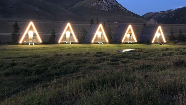A row of triangular tents with lights, set in a grassy field with mountains in the background.