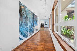 A hallway with wooden flooring, large abstract paintings on the walls, and a window with plants.