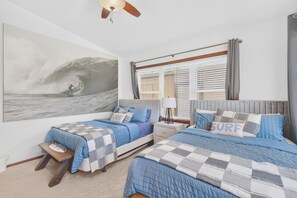 Une chambre avec deux lits, un grand tableau représentant une vague accroché au mur et un ventilateur de plafond.