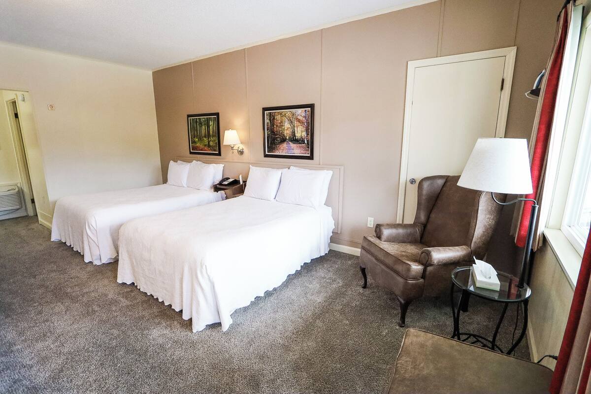Habitación de hotel con dos camas, un sillón de cuero marrón, una mesita de noche con lámpara y dos cuadros en la pared.