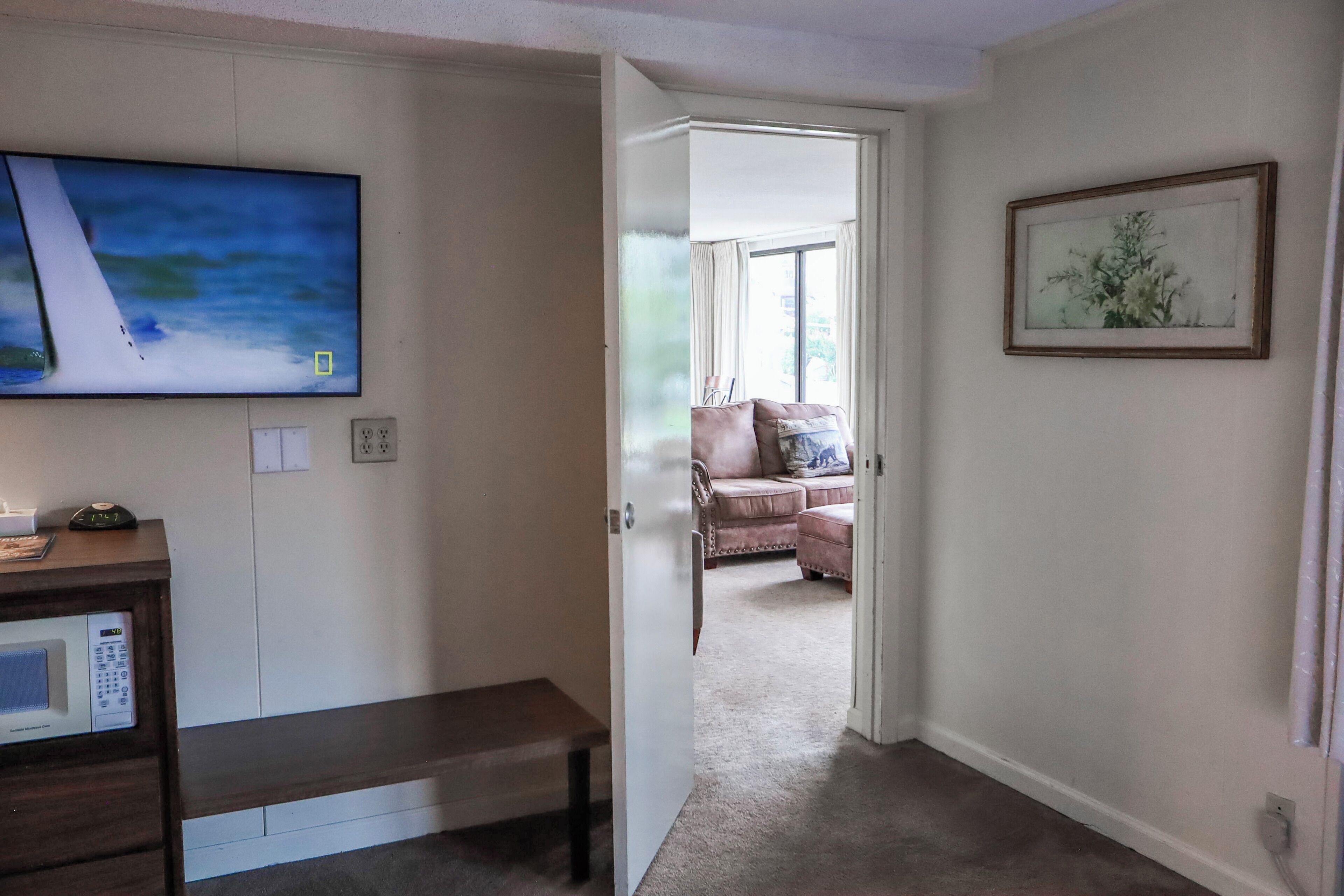 A living room with a flat-screen TV, a microwave, a bench, a door leading to another room, and a framed picture on the wall.