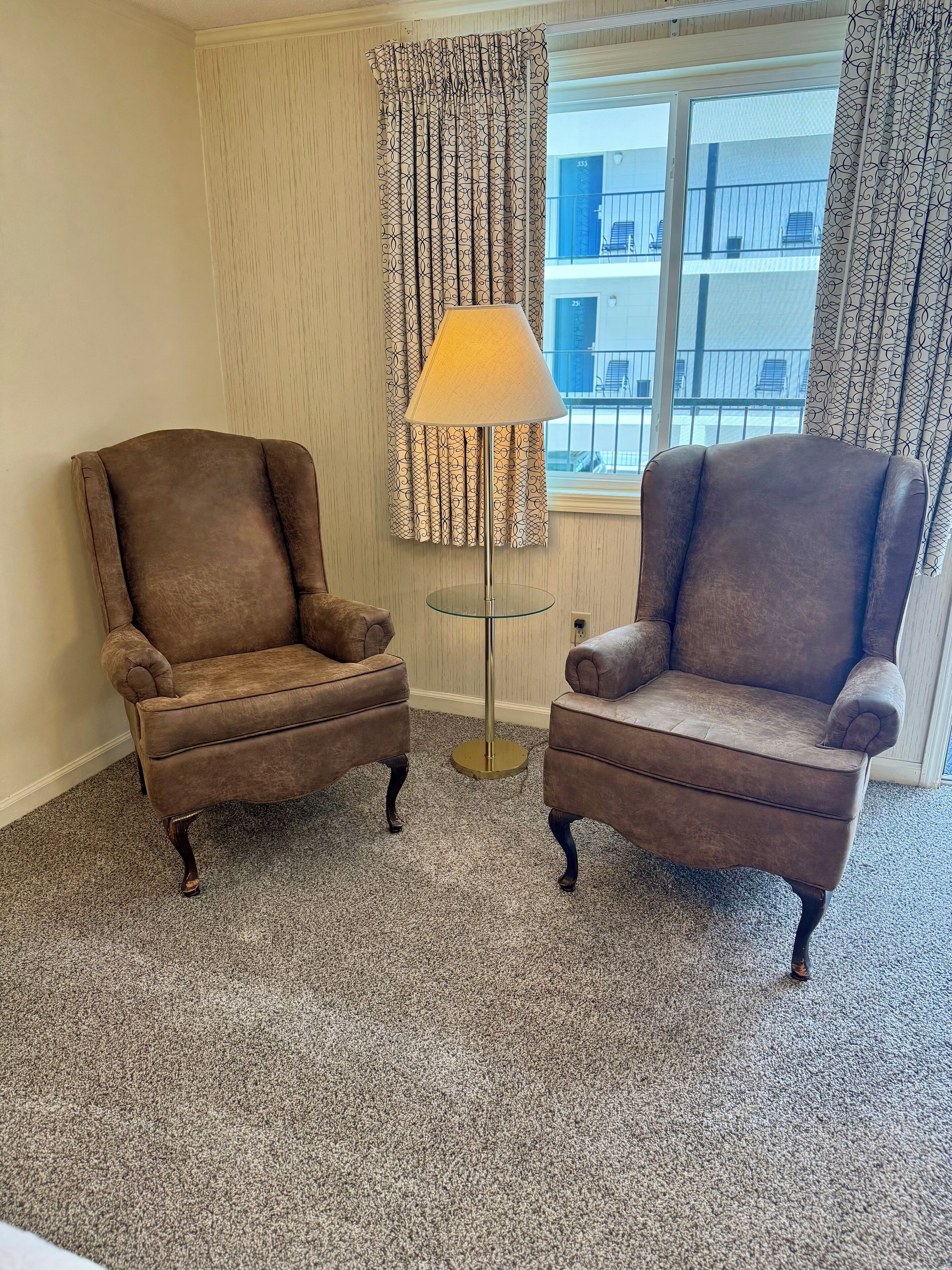 Two brown armchairs facing each other with a floor lamp between them, a patterned curtain, and a view of buildings through a window.