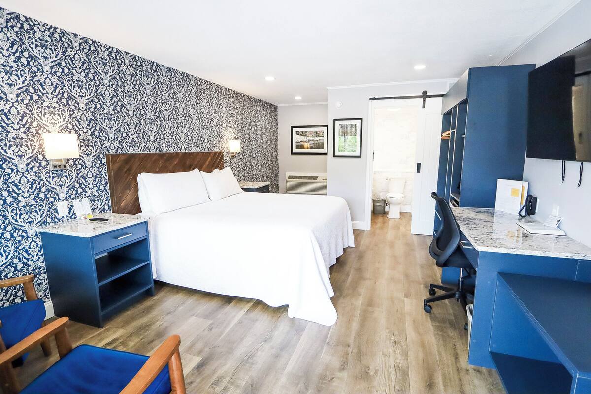 Una habitación de hotel moderna con una cama grande, un escritorio y una silla. Hay un televisor montado en la pared y una ventana que proporciona luz natural.