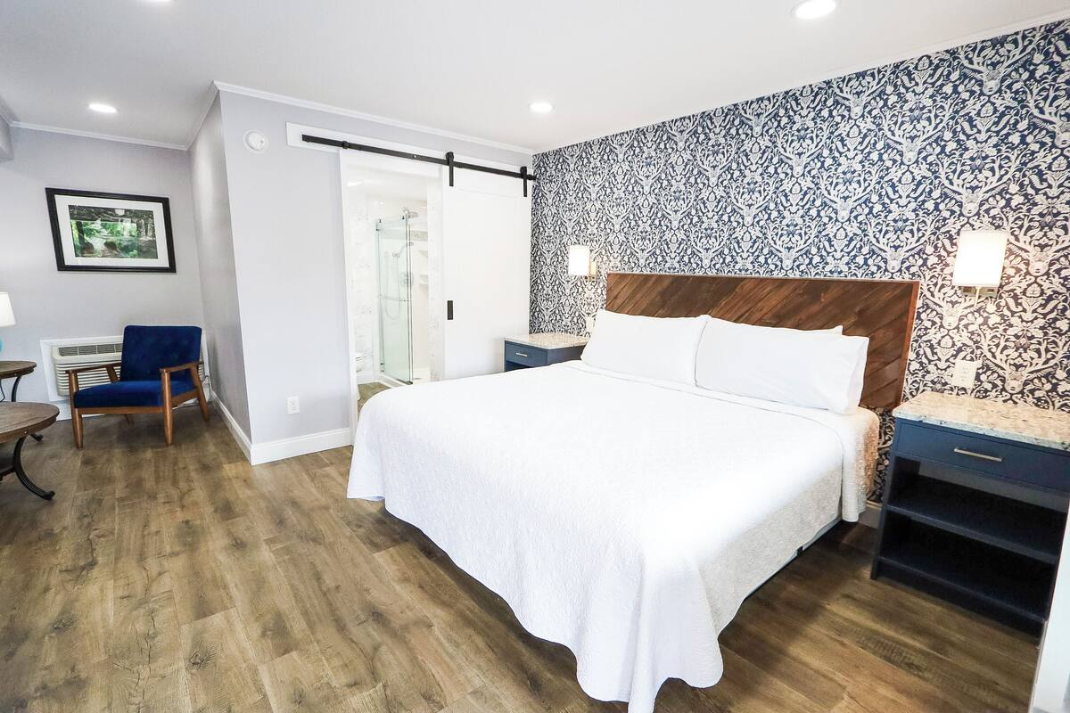 Una habitación de hotel moderna con una cama grande, suelo de madera, pared con diseño y un pequeño escritorio con lámpara.