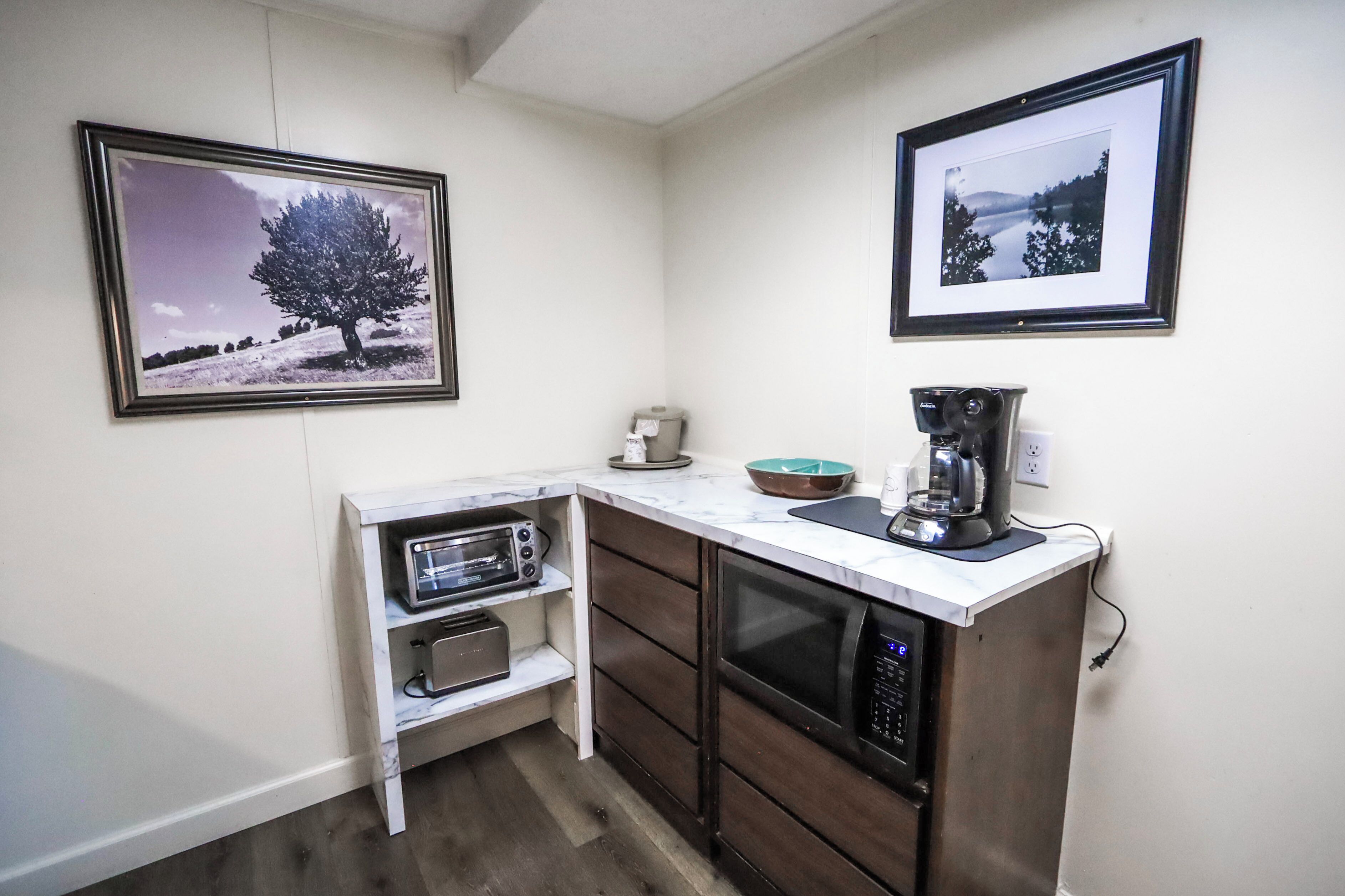 A compact kitchen with a microwave, toaster, and coffee maker on a white countertop. There are two framed pictures on the wall, one depicting a tree and the other a landscape.