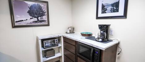 A compact kitchen with a microwave, toaster, and coffee maker on a white countertop. There are two framed pictures on the wall, one depicting a tree and the other a landscape.