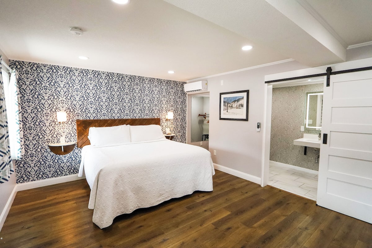 Un dormitorio con cama, papel tapiz estampado, suelo de madera, mesita de noche, apliques en la pared, una puerta y un baño visible a través de una puerta abierta.