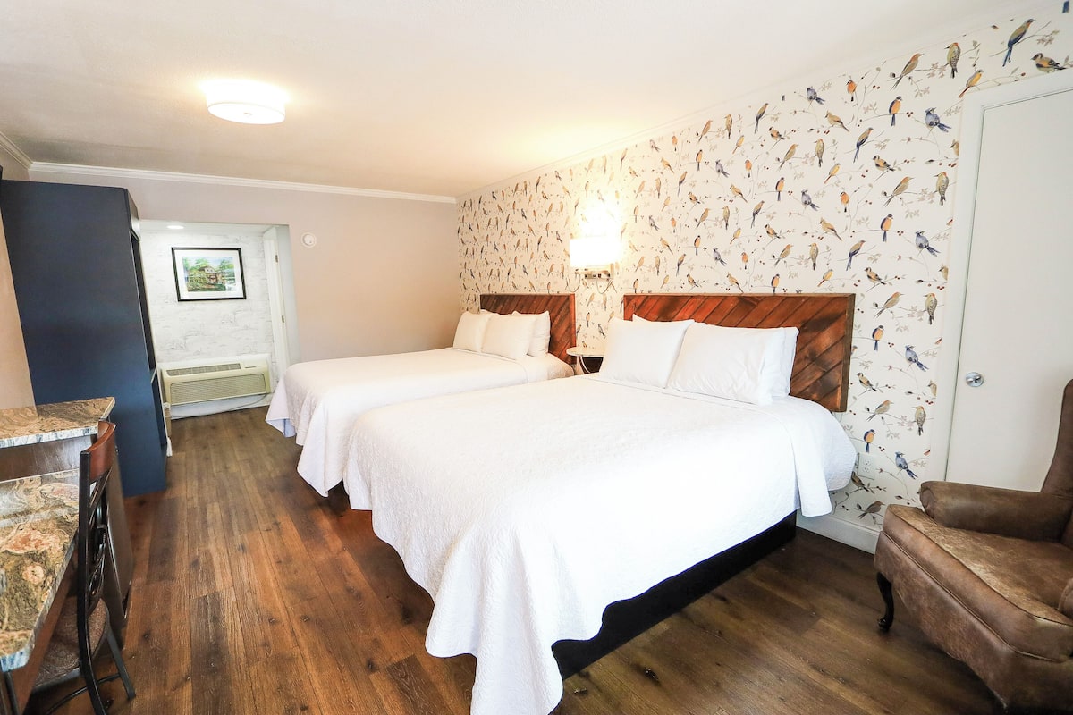 Habitación de hotel con dos camas, piso de madera, pared con papel tapiz de pájaros y una pequeña mesa con sillas.