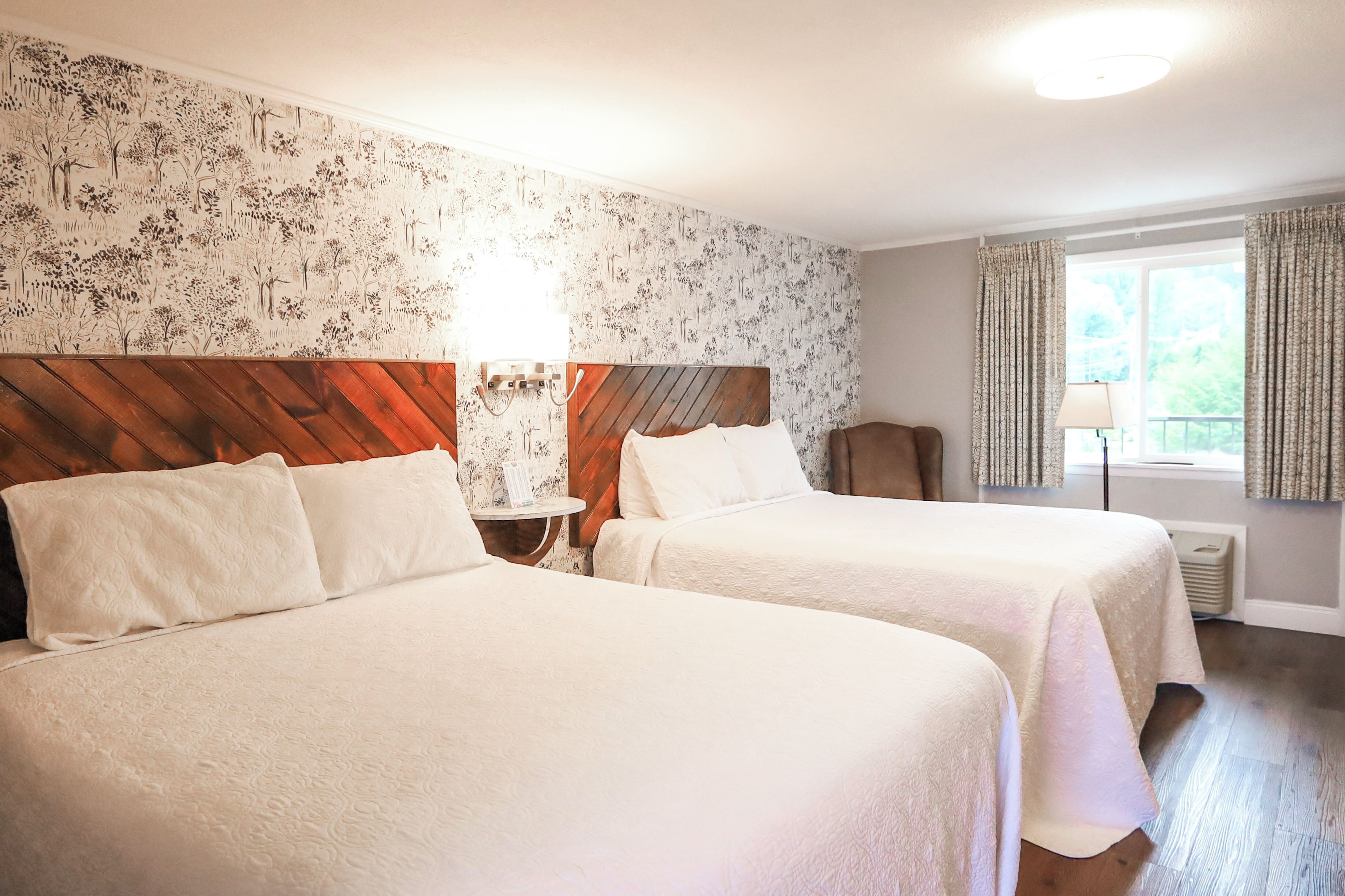 A hotel room with two beds, a wooden headboard, a nightstand, a lamp, and a window with curtains.
