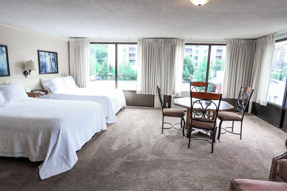 Habitación de hotel con dos camas, una mesa de comedor con sillas y ventanas amplias con cortinas.
