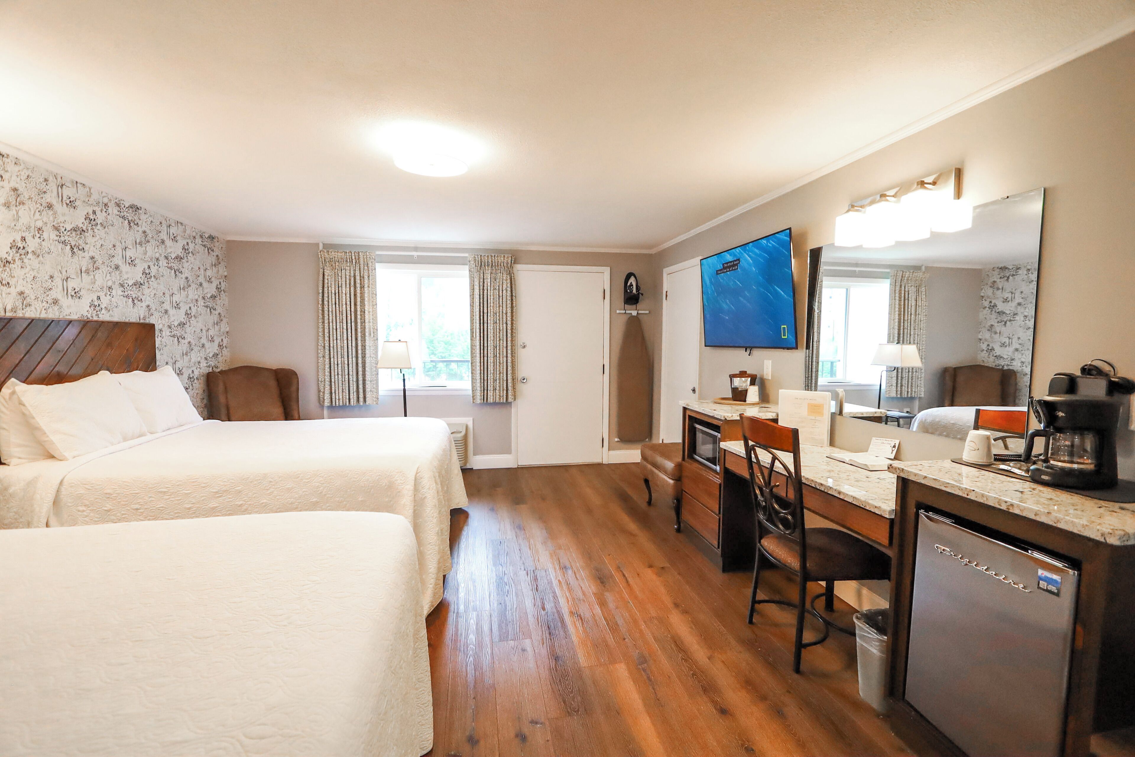 A hotel room with two beds, a desk with a chair, a refrigerator, a television, and a window with curtains.