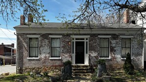 Ein zweistöckiges Backsteinhhaus mit Veranda, einer schwarzen Tür und einem kleinen Garten mit verschiedenen Pflanzen und Blumen.