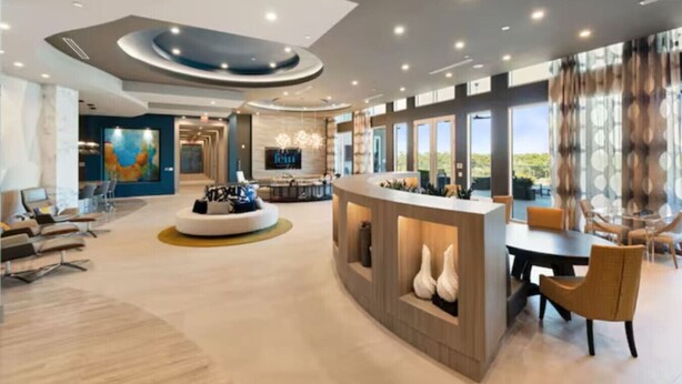 A spacious hotel lobby with a central seating area, a reception desk, and large windows offering a view of the outdoors.