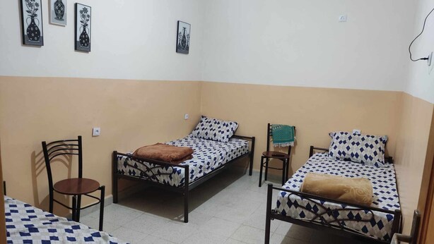 A dormitory room with two single beds, a chair, a small table, and wall art. - mirana mayotte hostel 47 (Ghardaia)
