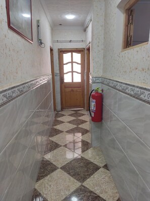 A narrow hallway with patterned tiles, a wooden door, and a fire extinguisher mounted on the wall. - mirana mayotte hostel 47 (Ghardaia)