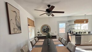Une salle à manger avec une longue table, des chaises, un ventilateur de plafond et une cuisine en arrière-plan.