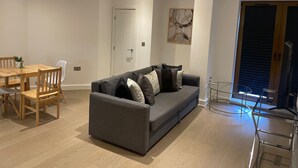 A modern living room with a grey sofa, wooden dining table, and chairs. There is a door, a painting on the wall, and a TV stand with a TV. - Spacious 1 Bedroom Apartment in St Albans (St Albans)