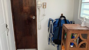 Een kamer met een houten deur, een vloer betegeld met witte tegels, een klein houten tafeltje met een fles, een blauwe tas en een plank met opgevouwen handdoeken.