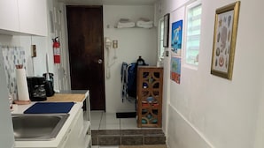 Een smalle gang met een keukenhoek aan de linkerkant, met een spoelbak, kastjes en een koffieapparaat. Een houten deur en een brandblusser zijn zichtbaar aan de muur. De gang heeft een betegelde vloer met een vloerkleed dat naar een trap leidt.